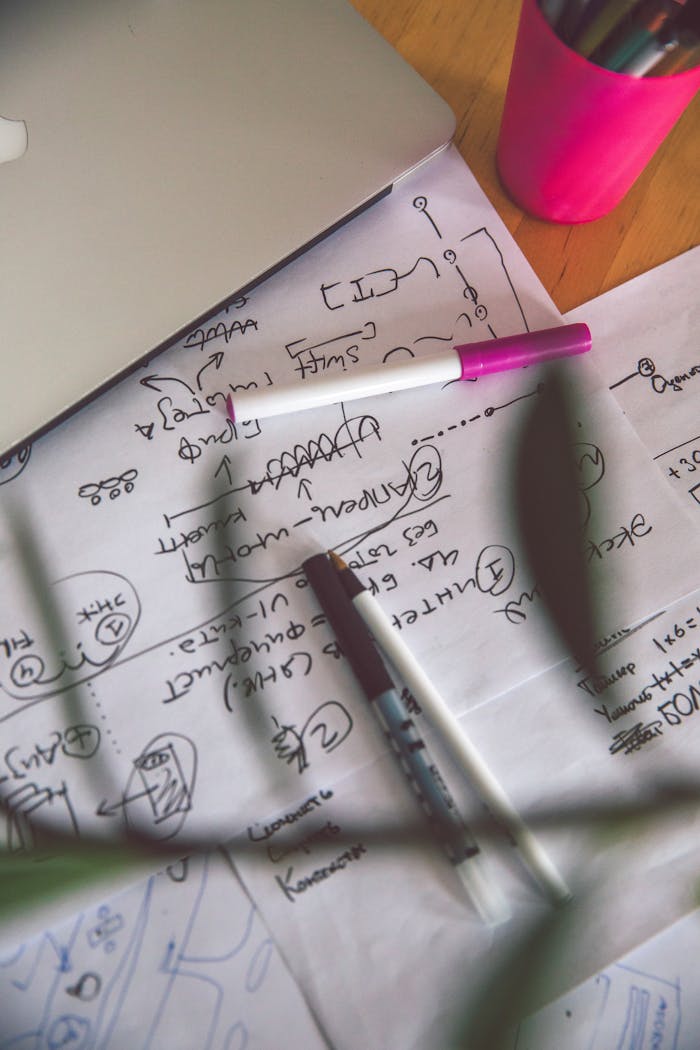 A top view of a creative workspace with a laptop, pens, and hand-drawn sketches on paper.